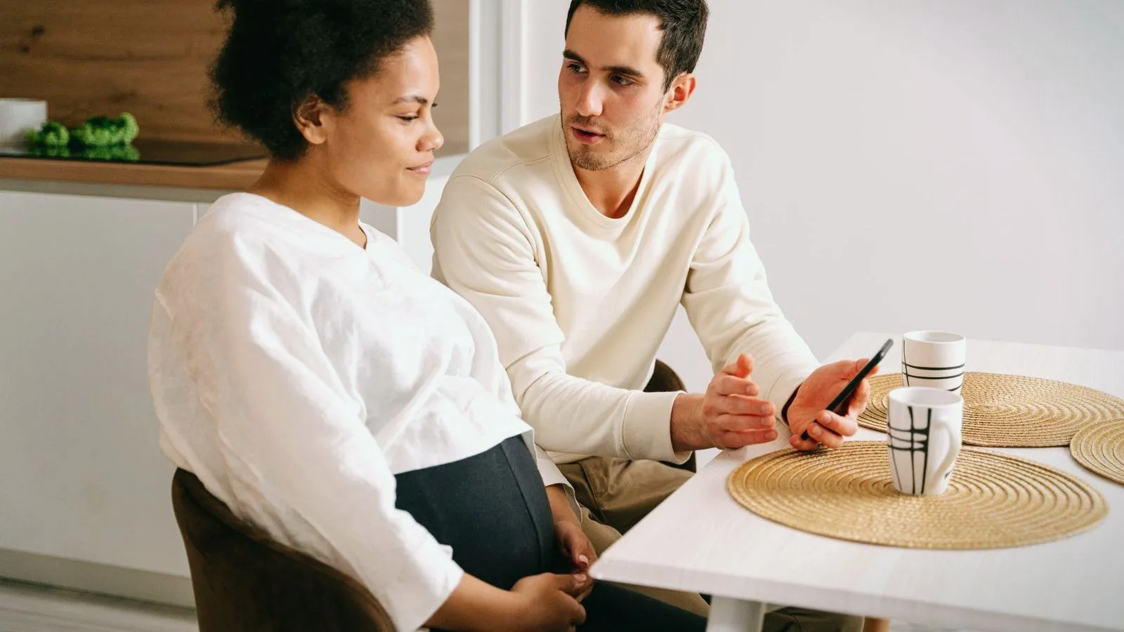 A pregnant person and a partner sitting together at a kitchen table with mugs and a phone