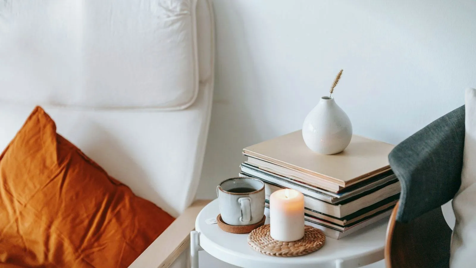 A small lit candle and a mug on a side table stacked with books, next to a chair with an orange cushion