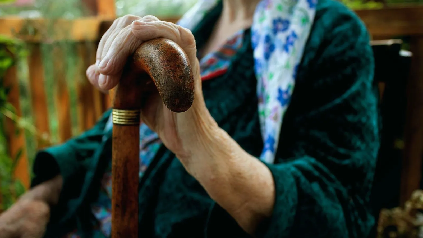 An older person's hand rests on the curved handle of a wooden cane, in front of a dark green jacket and a blue floral scarf.