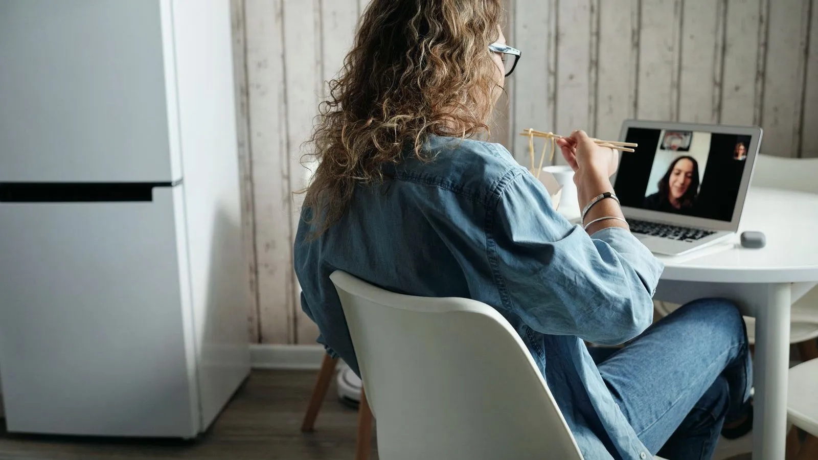 A woman seen from behind at a kitchen table eats with chopsticks while on a video call on a laptop.