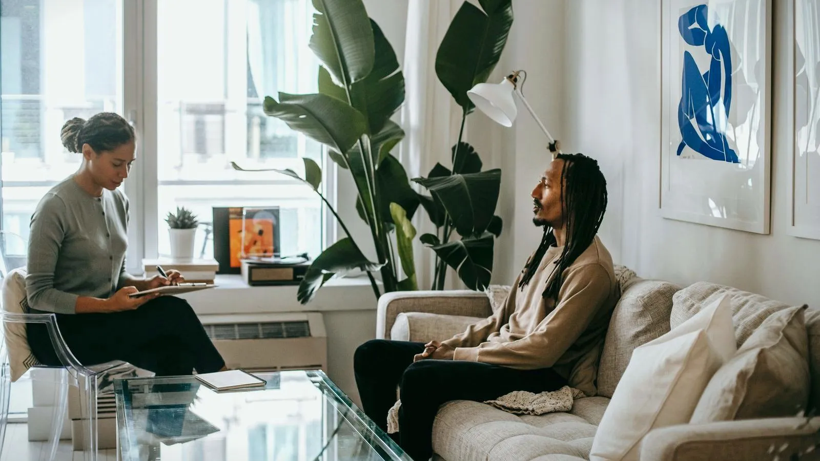 A therapist taking notes while talking with a client seated on a sofa in a bright room.