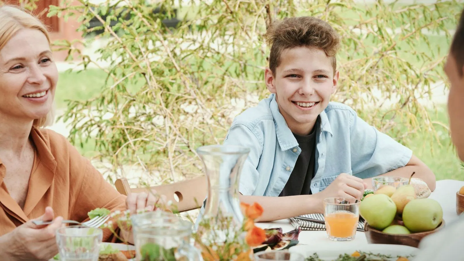 A smiling boy and an adult sharing a meal at an outdoor table with fruit and juice.