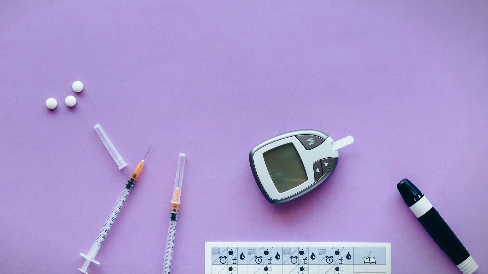 A blood glucose meter, two syringes, tablets and a lancet pen laid out on a purple background