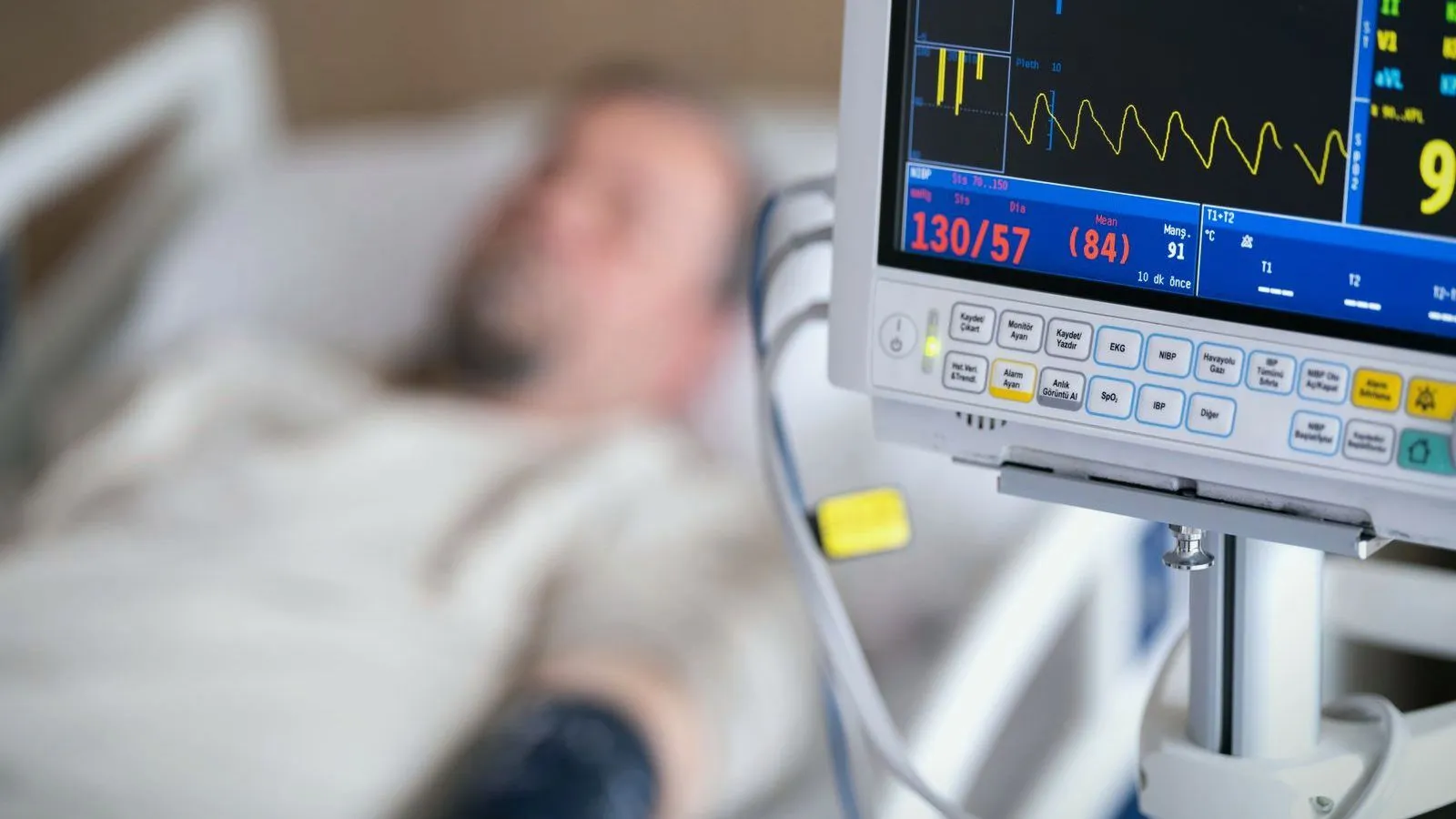 A bedside monitor showing vital signs next to a patient lying out of focus in a hospital bed