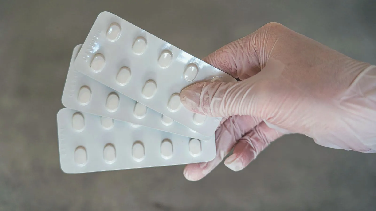 A gloved hand holding three blister strips of plain white tablets