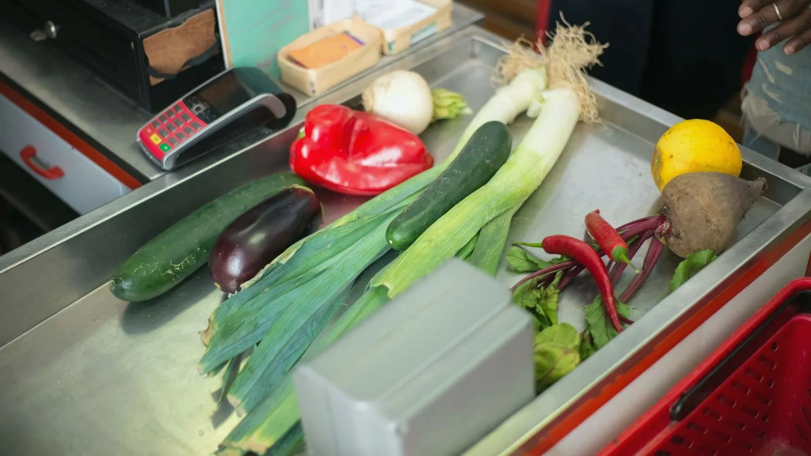 Fresh vegetables on a supermarket checkout counter beside a card reader and a red basket