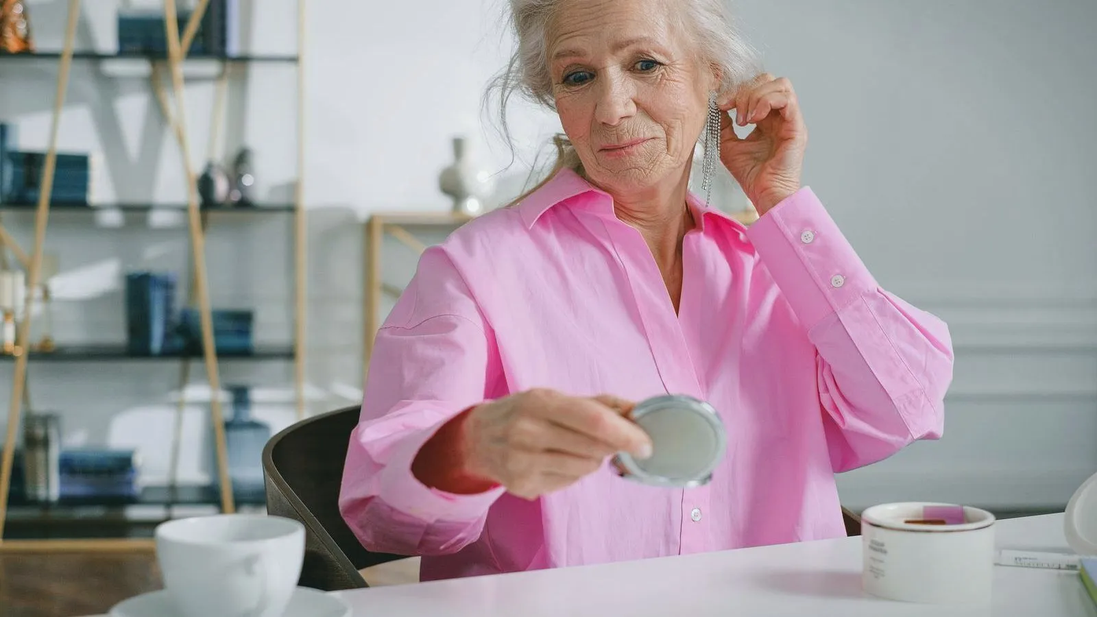 An older person fitting a hearing aid while sitting at a table with a cup