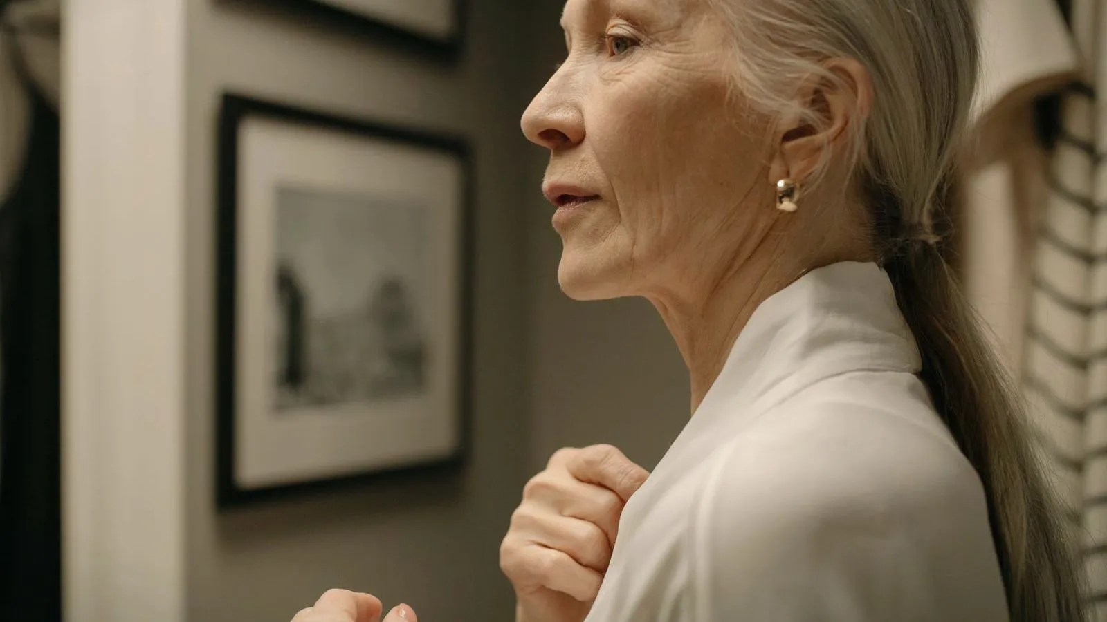 An older woman with long gray hair tied back, seen in profile with one ear visible, standing near framed pictures.