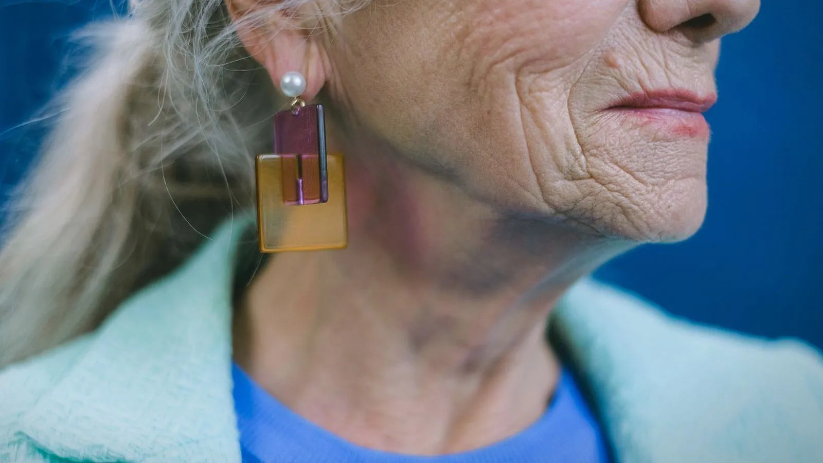 Close view of an older woman's ear and jaw, with grey hair tied back and a large drop earring.