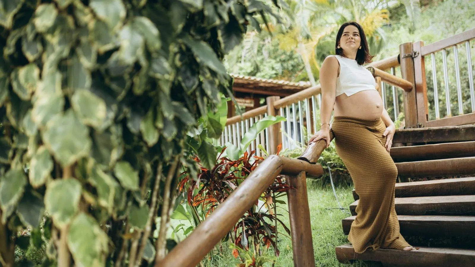 A pregnant woman leaning on a wooden railing outdoors among tropical plants.