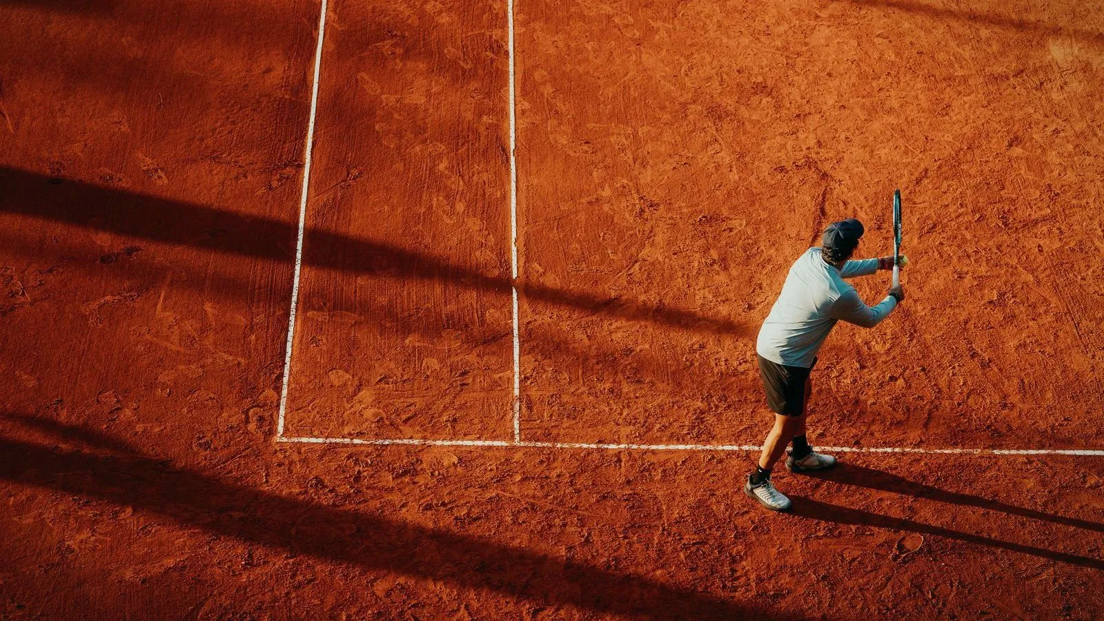 A tennis player preparing to serve on a sunlit clay court, seen from above.