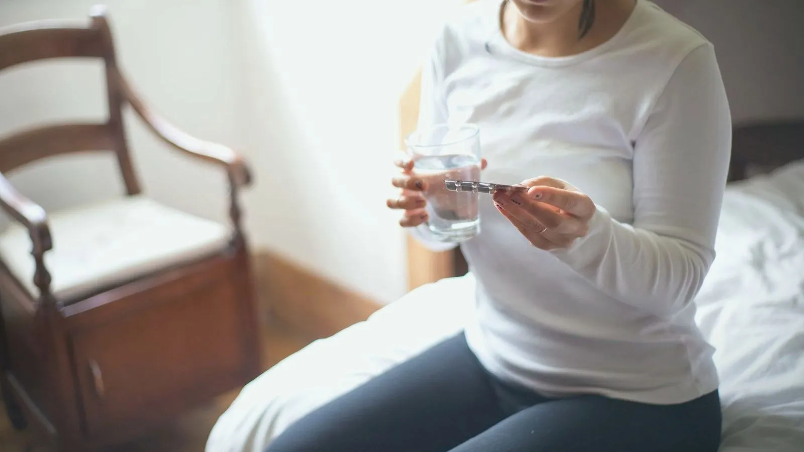 A person in a white top sits on the edge of a bed holding a glass of water and a strip of tablets, face out of frame.