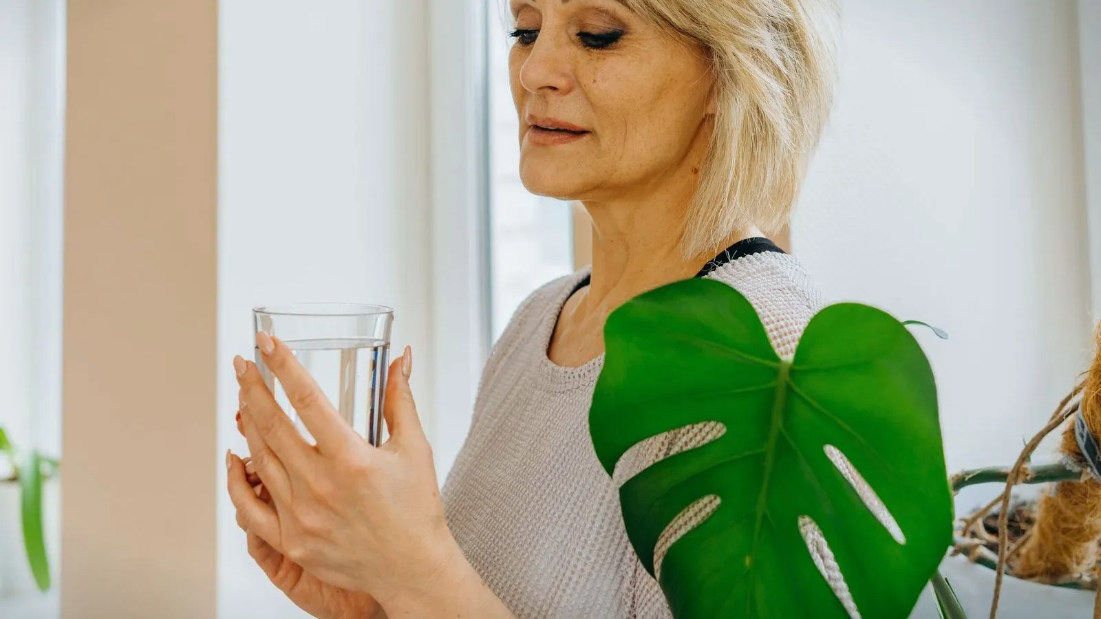 A woman in her fifties holding a glass of water beside a window, a large green leaf in the foreground