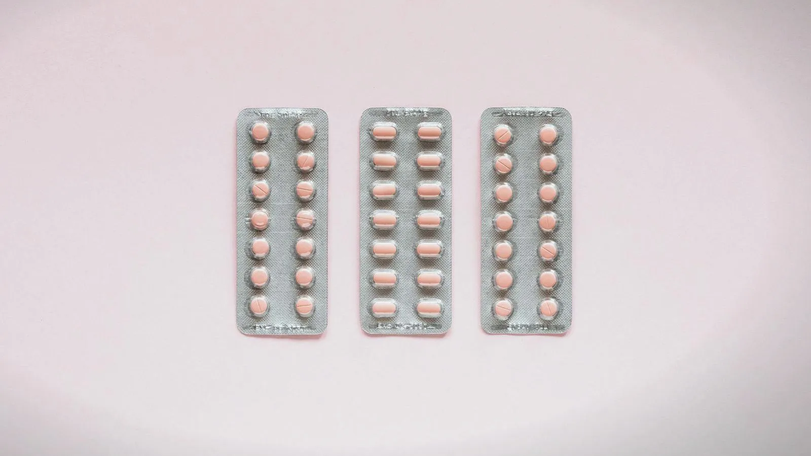 Three blister strips of pale pink tablets laid side by side on a soft pink background.