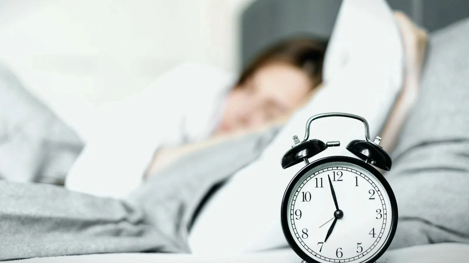 A person asleep in bed behind a black alarm clock.