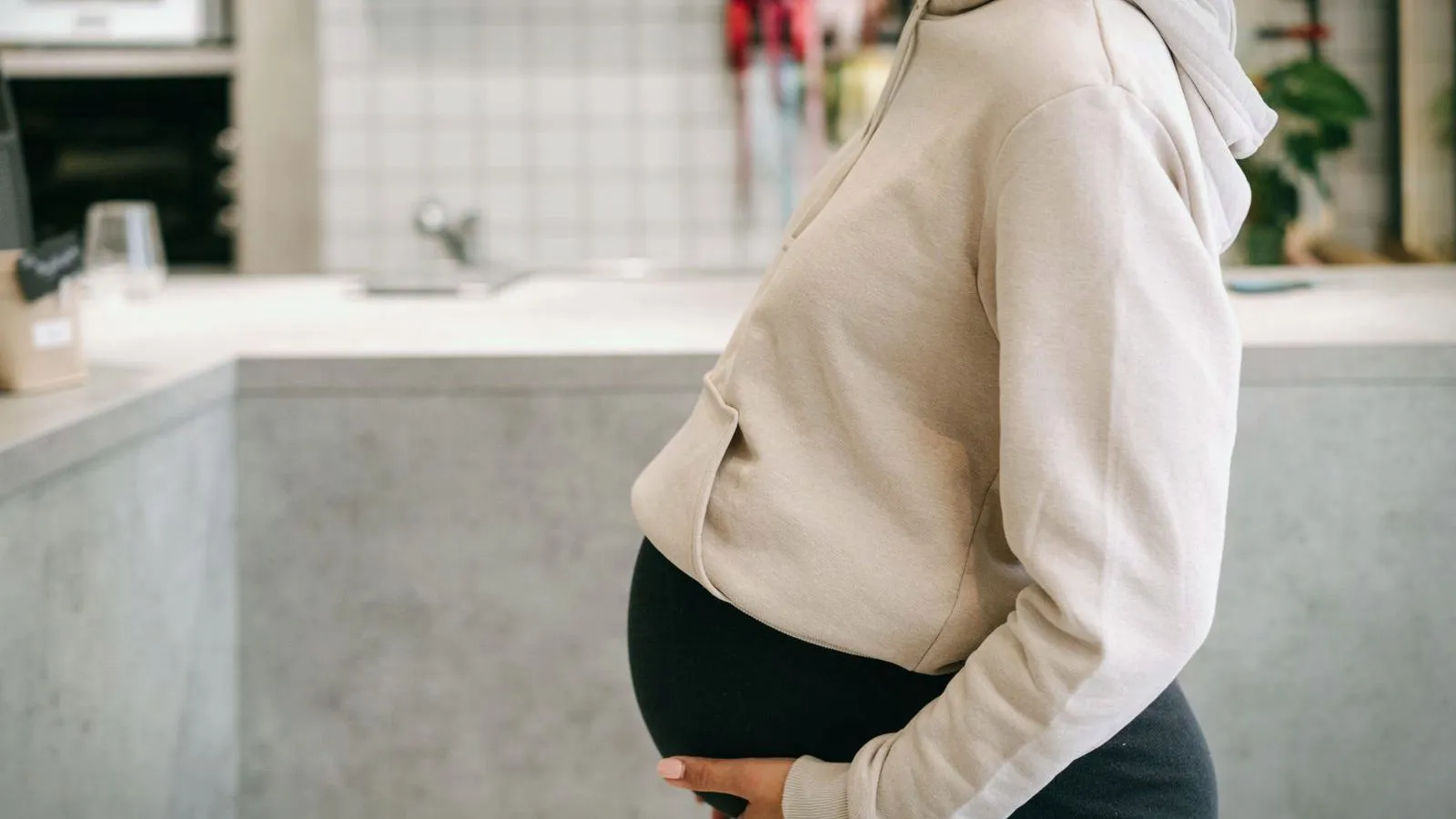 A pregnant person in a pale hoodie stands side-on in a kitchen with a hand under their belly, face out of frame.