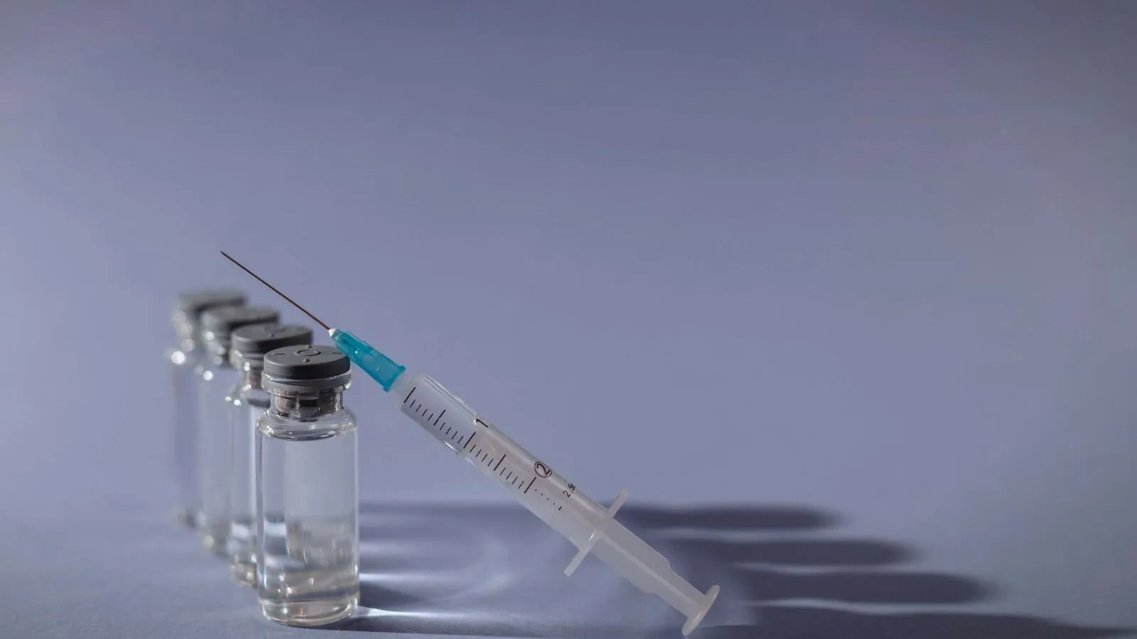 A row of clear glass vials with a syringe leaning against them on a gray-violet surface.