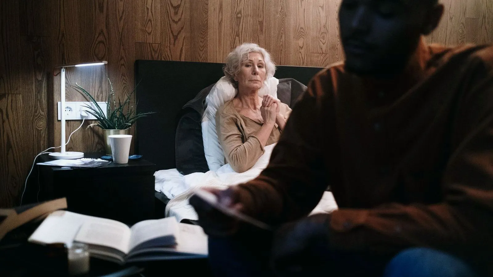 An older woman with white hair sitting up in bed with hands clasped, while a second person sits in shadow in the foreground.
