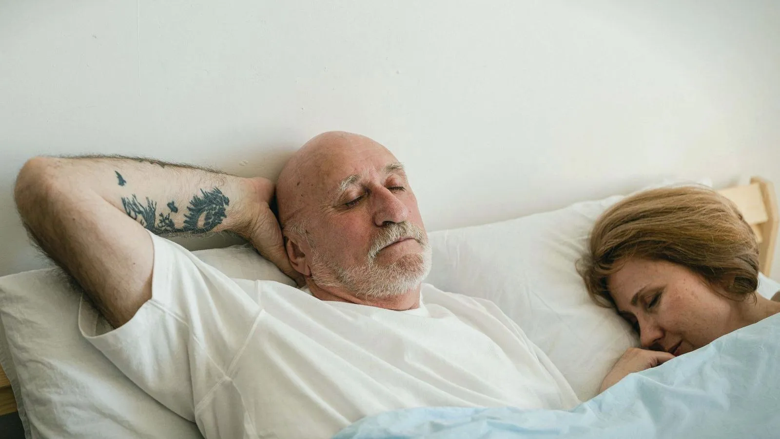 An older man lying awake in bed with his arms behind his head while a woman sleeps beside him