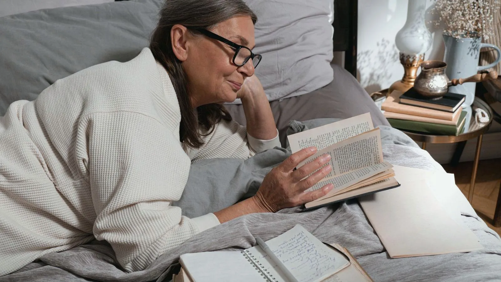 An older woman in glasses lying in bed reading a book.