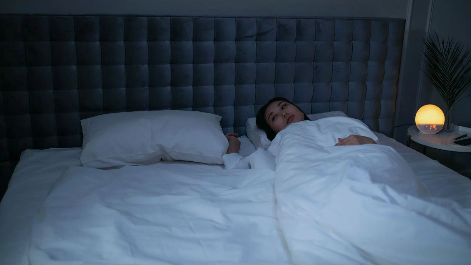 A woman lying awake in a large bed in a dark bedroom, a small lamp lit on the nightstand