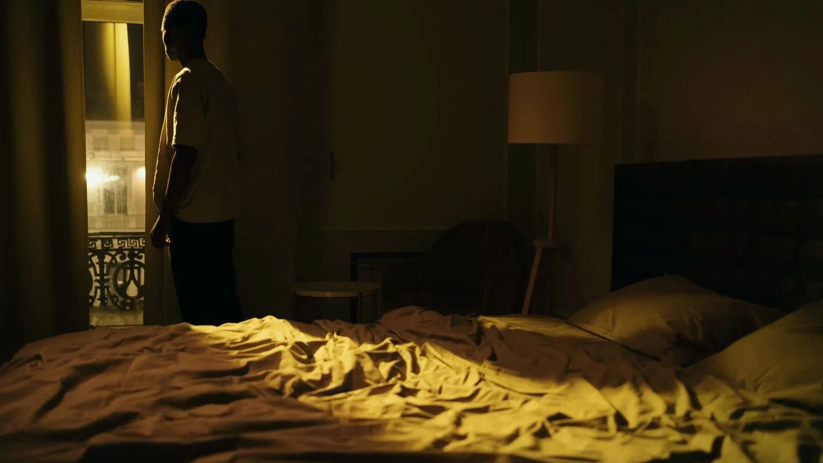A dark bedroom at night with a person standing in silhouette by a lit doorway and an unmade bed in the foreground.