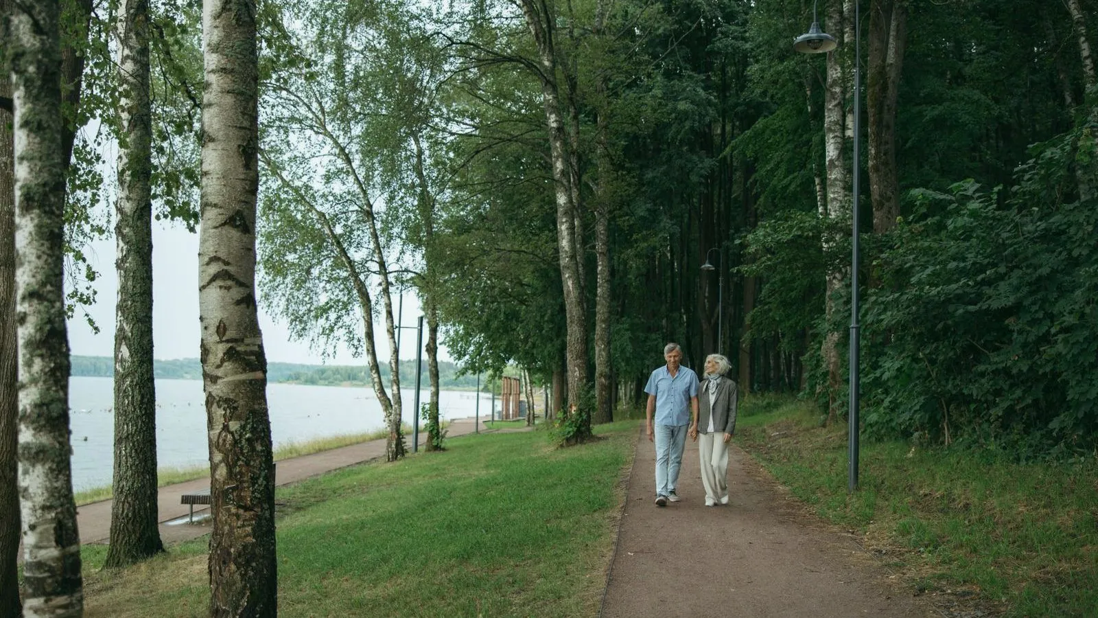 An older couple walks along a tree-lined path beside a lake.