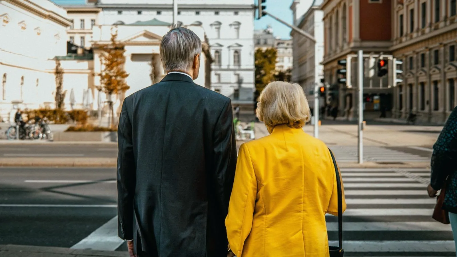 An older couple seen from behind stand at a pedestrian crossing on a sunny city street lined with old buildings.