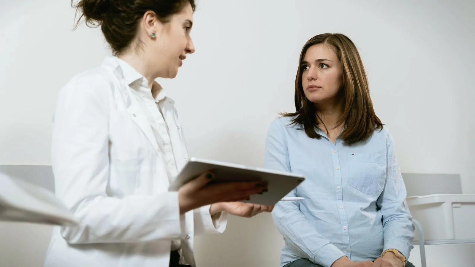 A doctor holding a tablet talks with a young woman in a clinic consultation room