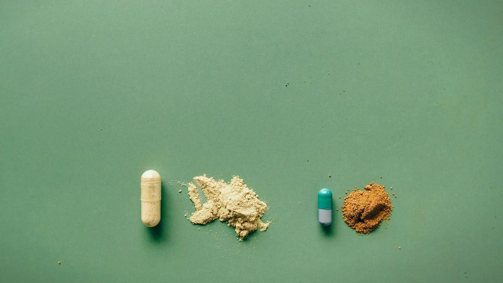 A beige capsule beside a small heap of beige powder, and a green and white capsule beside a heap of brown powder, on a green surface.