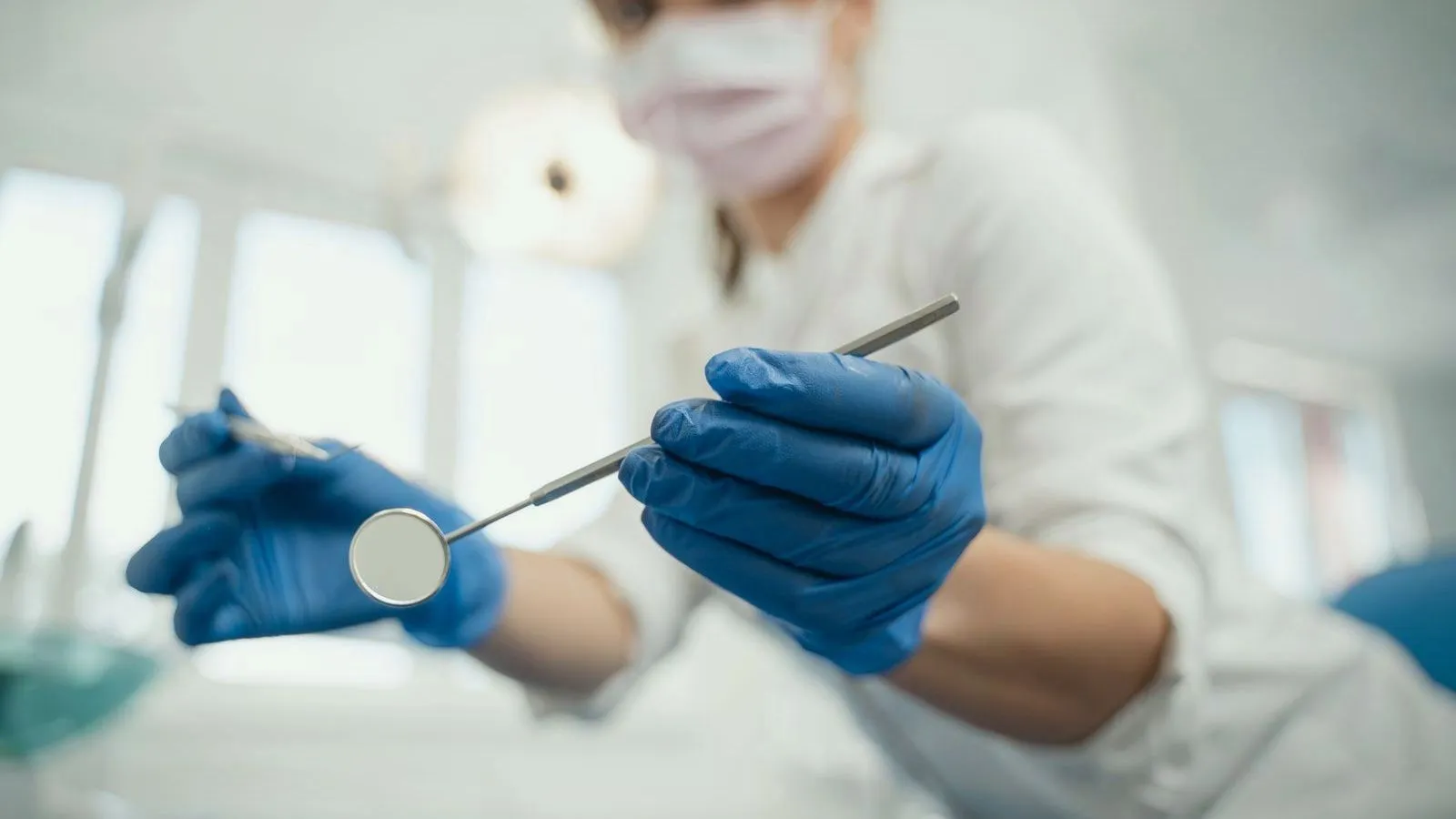A masked dentist in blue gloves holds a dental mirror and probe, seen from the patient's chair.