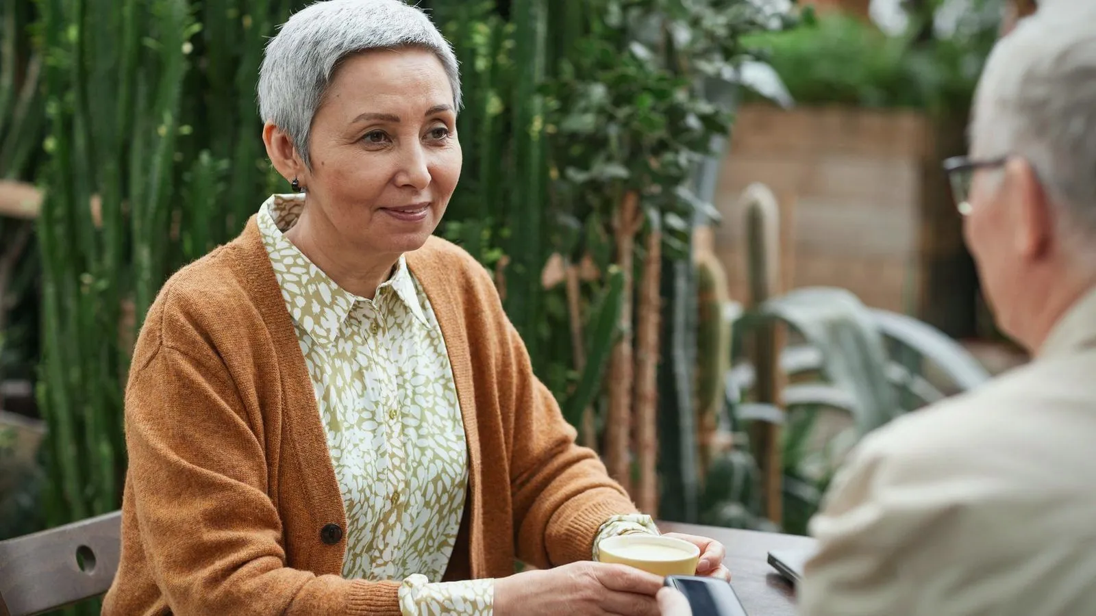 An older woman talking with a companion over coffee among green plants