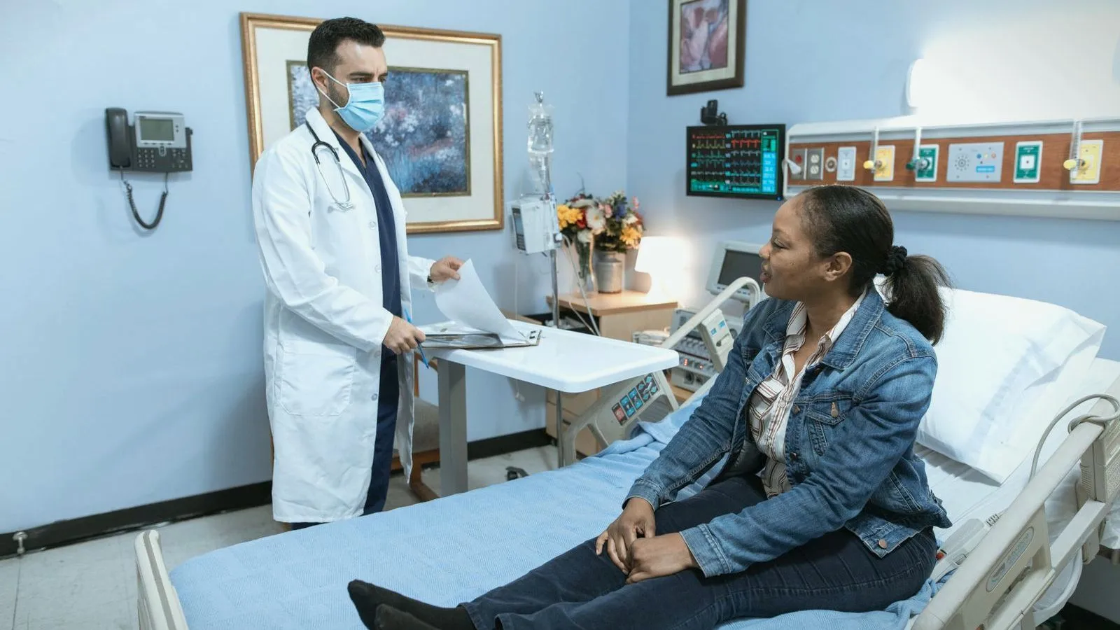 A doctor in a white coat and mask talks with a woman sitting on a hospital bed