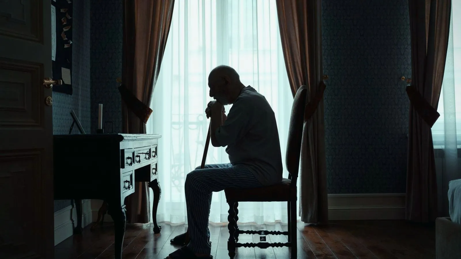 A person sitting alone on a chair beside a curtained window in a dim room
