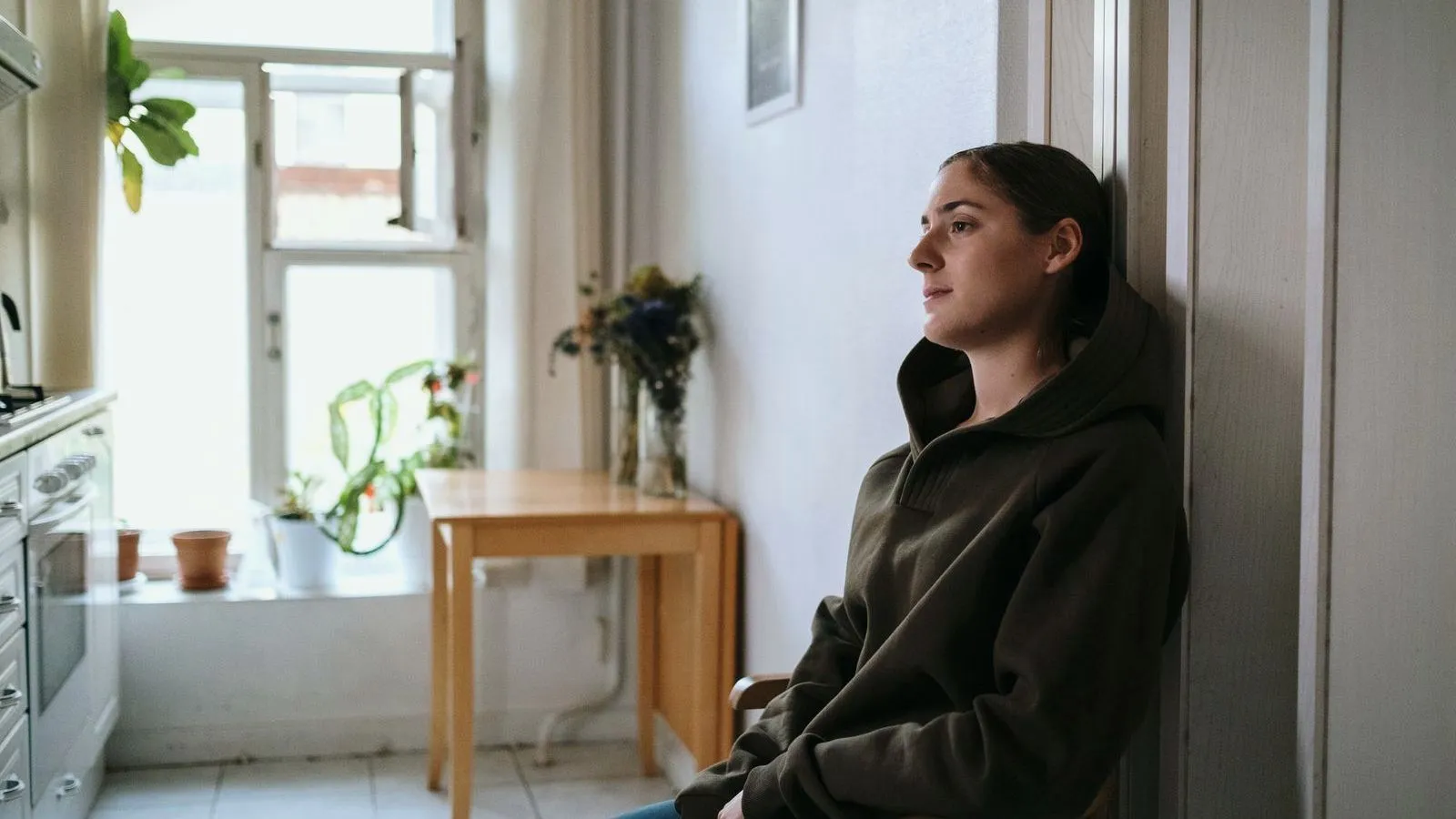 A woman sitting alone on a kitchen chair, looking towards a window