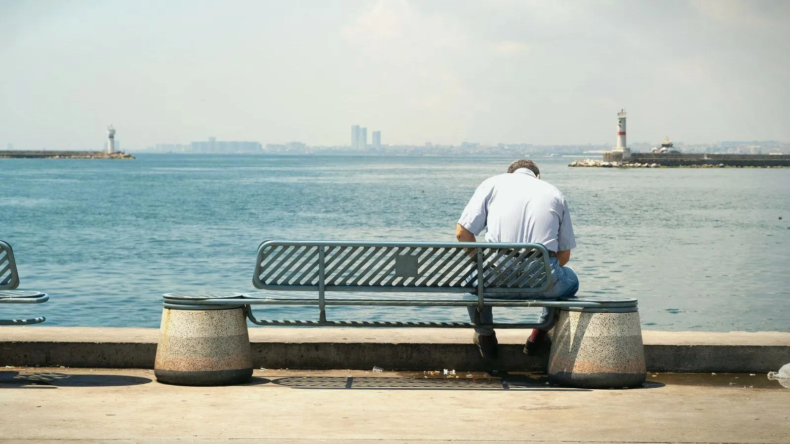A person in a white shirt, seen from behind, sitting alone on a bench facing the sea, with lighthouses in the distance.