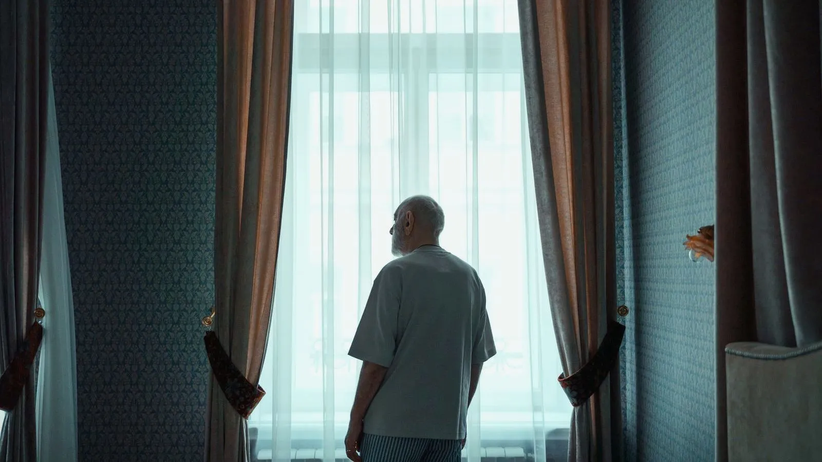 An older person standing alone at a tall curtained window, seen from behind