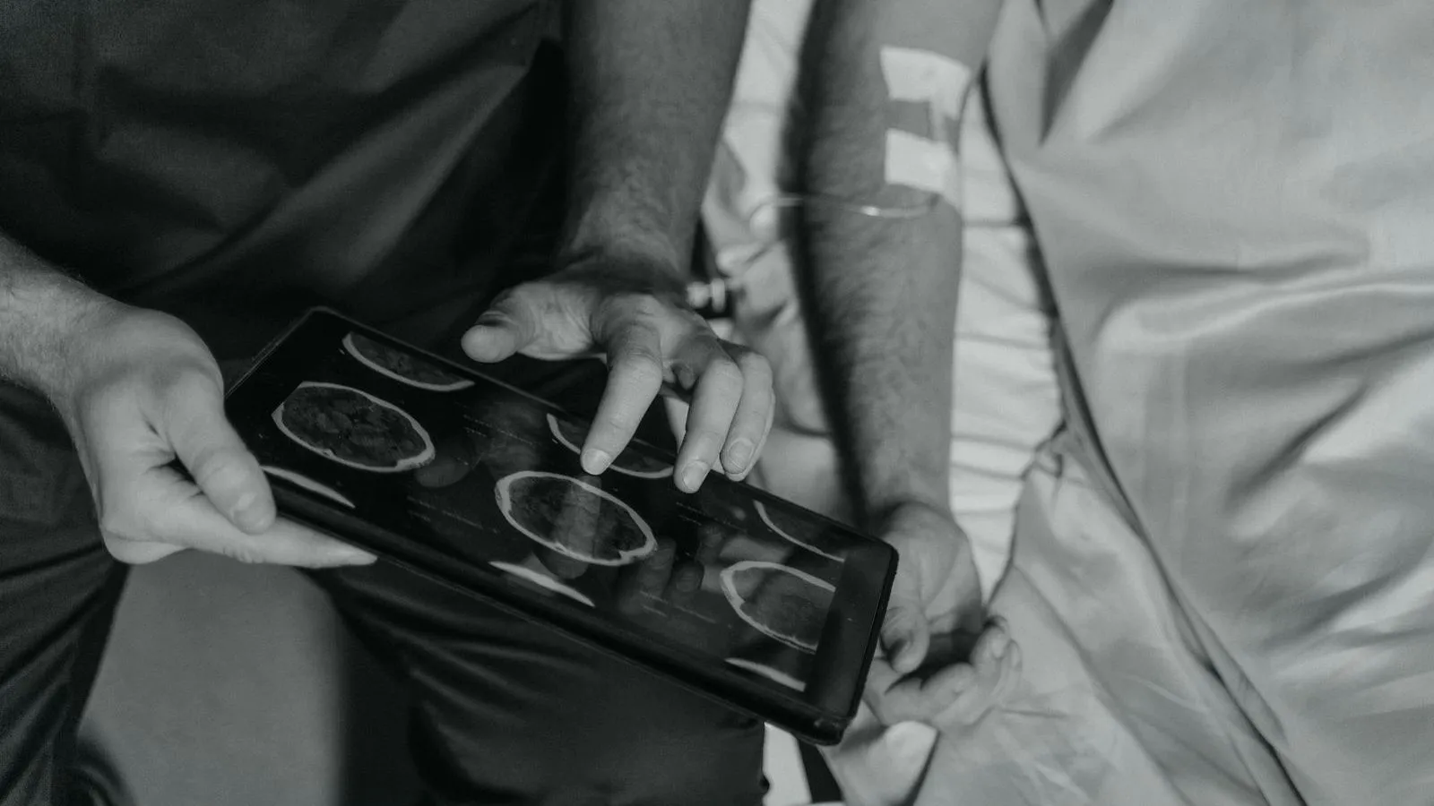 A clinician showing brain scan images on a tablet to a patient at a bedside, in black and white