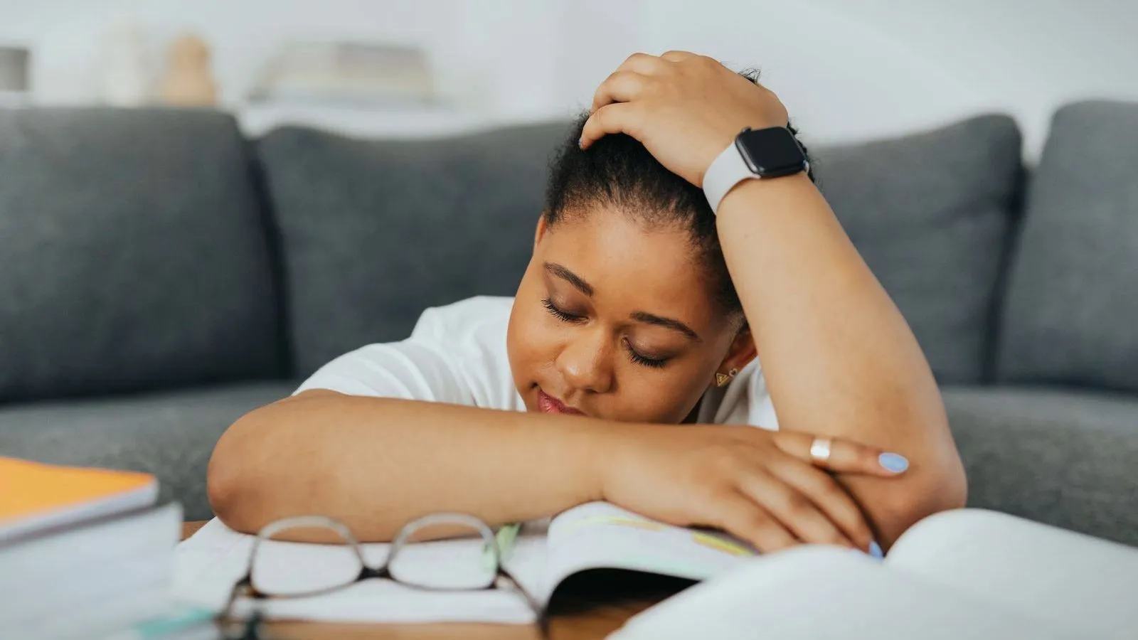 A person rests their head on a folded arm over an open book, eyes closed, with glasses set down beside them.