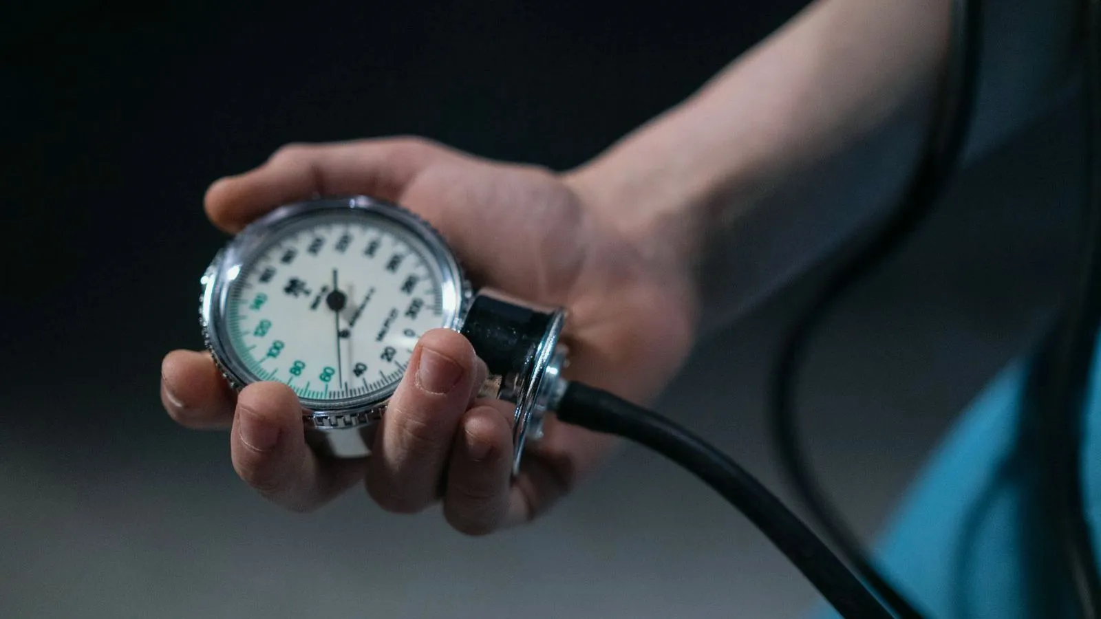 Close-up of a hand holding the round dial gauge of a manual blood pressure cuff, its black tubing trailing off.