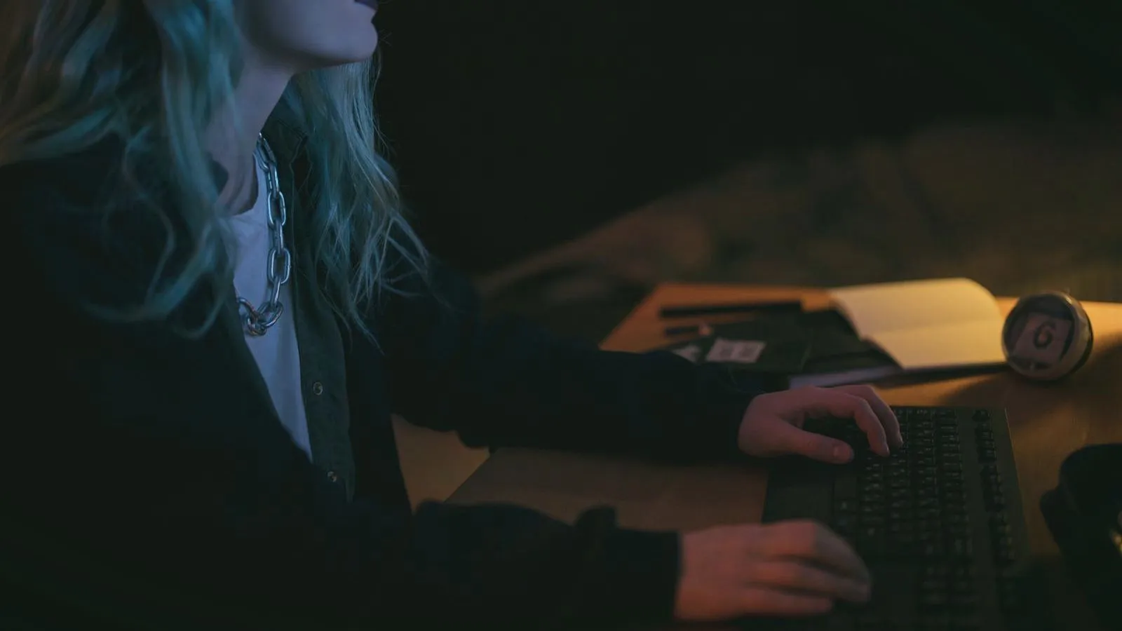 A person with long hair typing on a keyboard at a desk in a dark room, face out of frame.