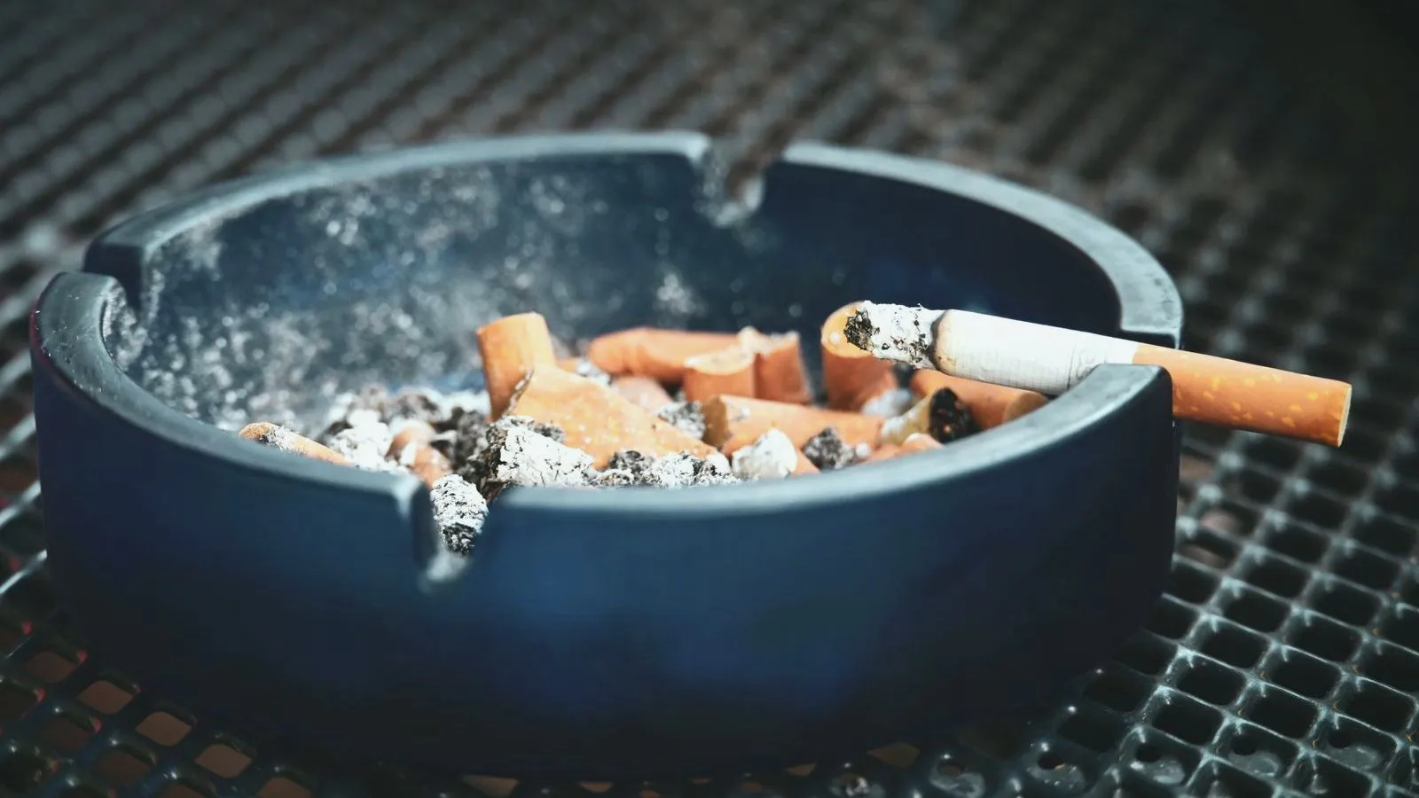 A glass ashtray of spent cigarette butts with one still burning