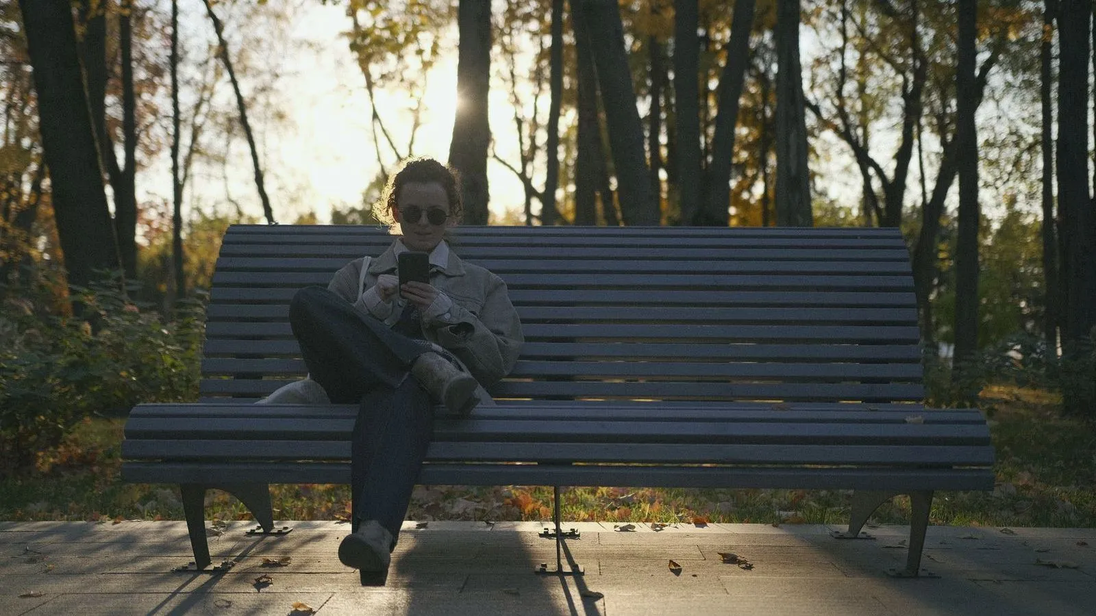 A person in sunglasses sits cross-legged on a wide park bench looking at a phone, with low sun coming through the trees behind.