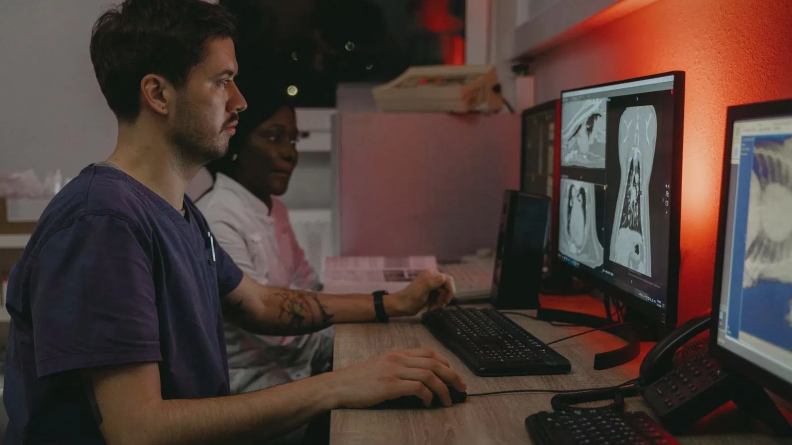 Two hospital staff sit at a desk with keyboards, looking at monitors that show black and white chest scan images.