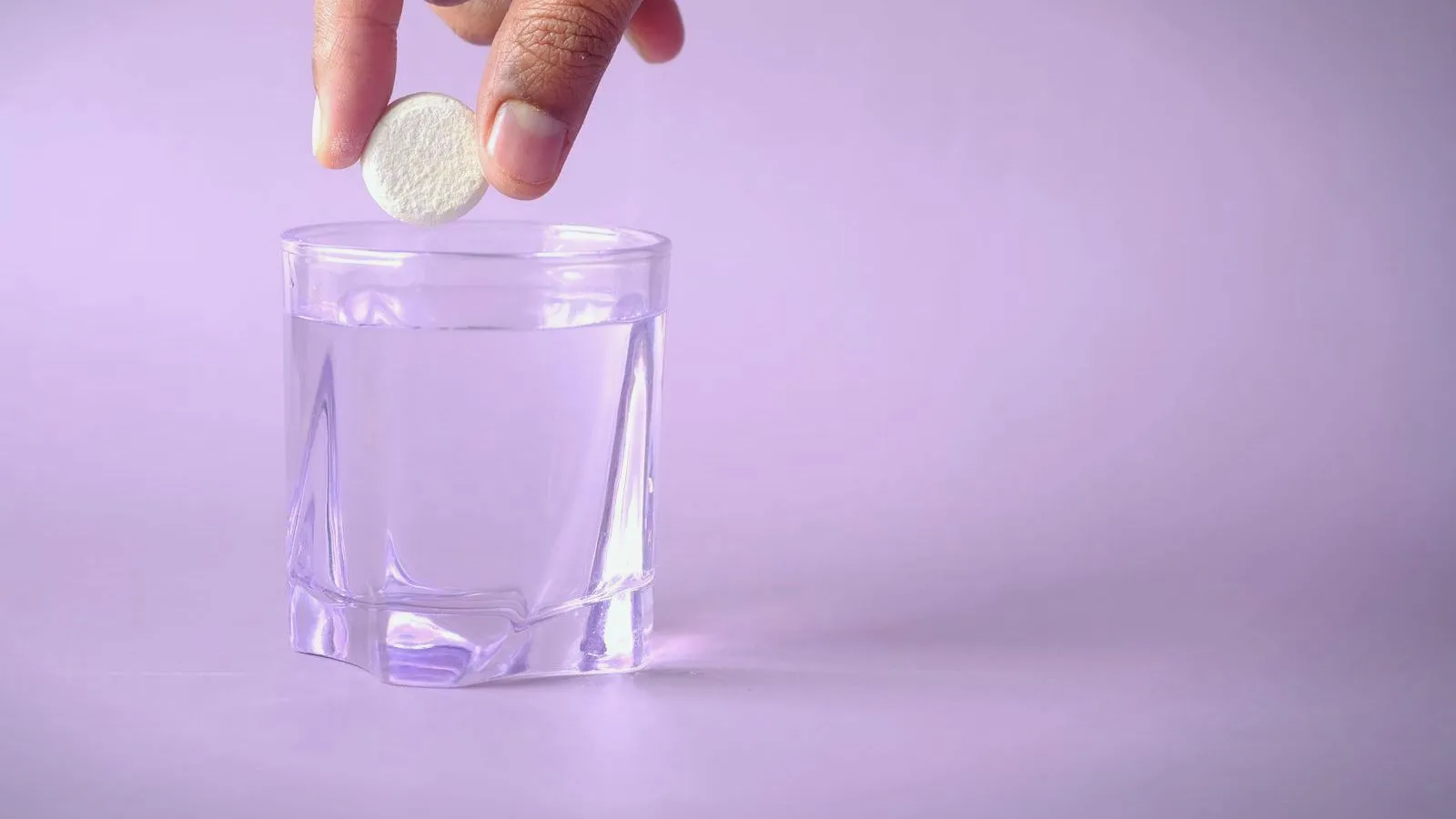 A hand dropping a white tablet into a glass of water against a lavender background.