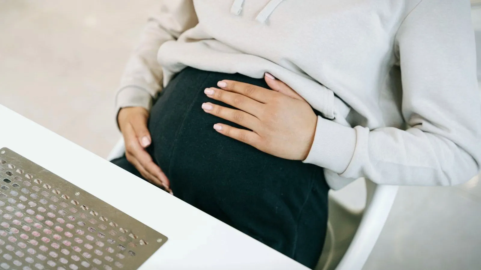 A pregnant woman in a cream sweater seated at a white desk with both hands resting on the belly, face not shown.