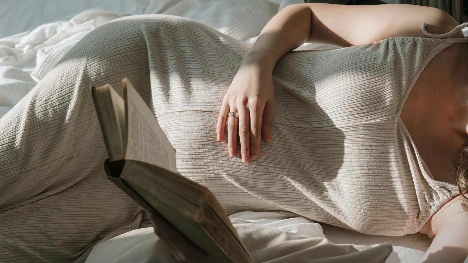 A pregnant person lying on a bed in a ribbed dress, one hand resting on the belly and an open book in front, face out of frame.