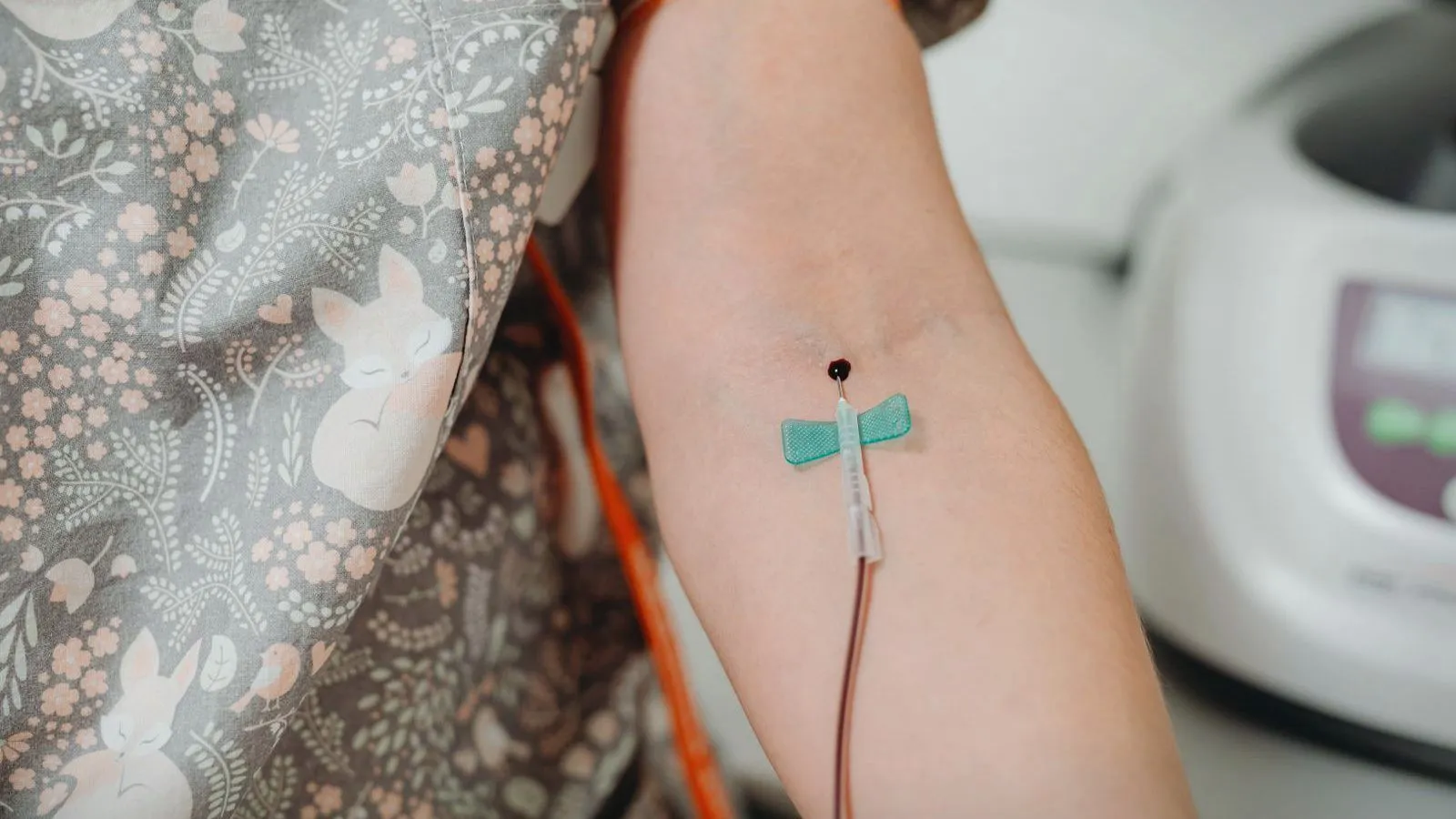 A blood sample being drawn from a person's arm through a winged needle, with a laboratory machine behind