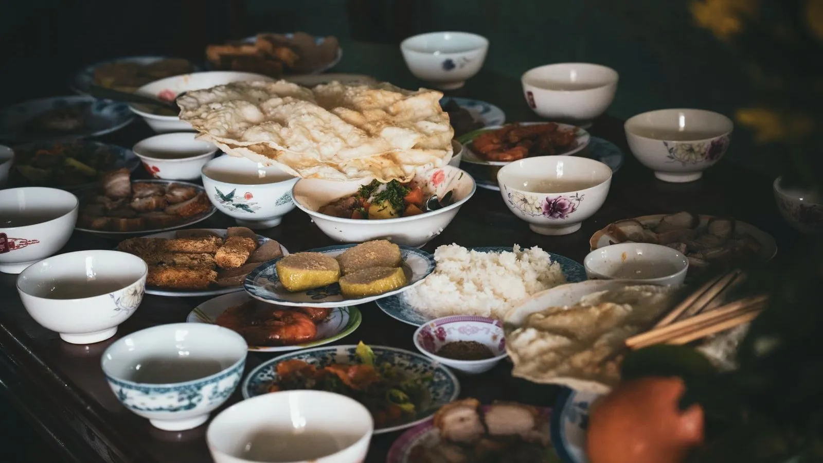 A dark table crowded with small bowls and plates of rice, soup and side dishes, teacups around the edge