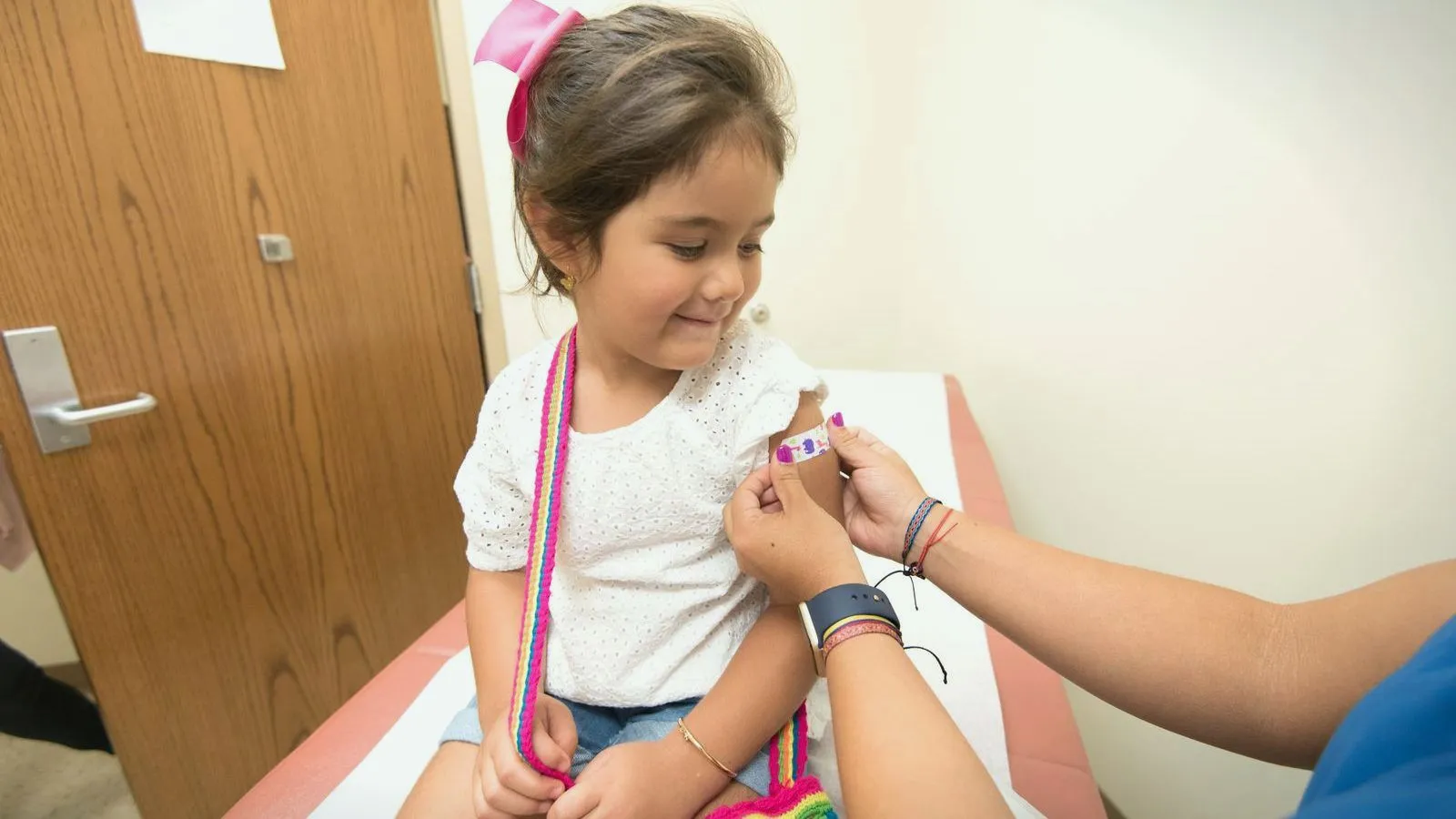 A smiling young girl sits on an exam table while an adult presses a small bandage onto her upper arm.